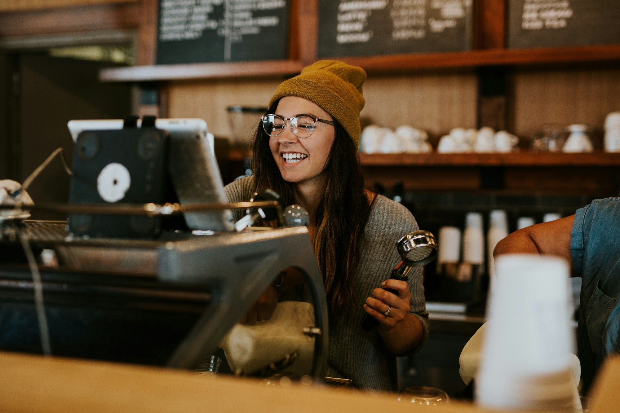 A smiling barista in a mustard beanie and glasses working at the counter of a busy café