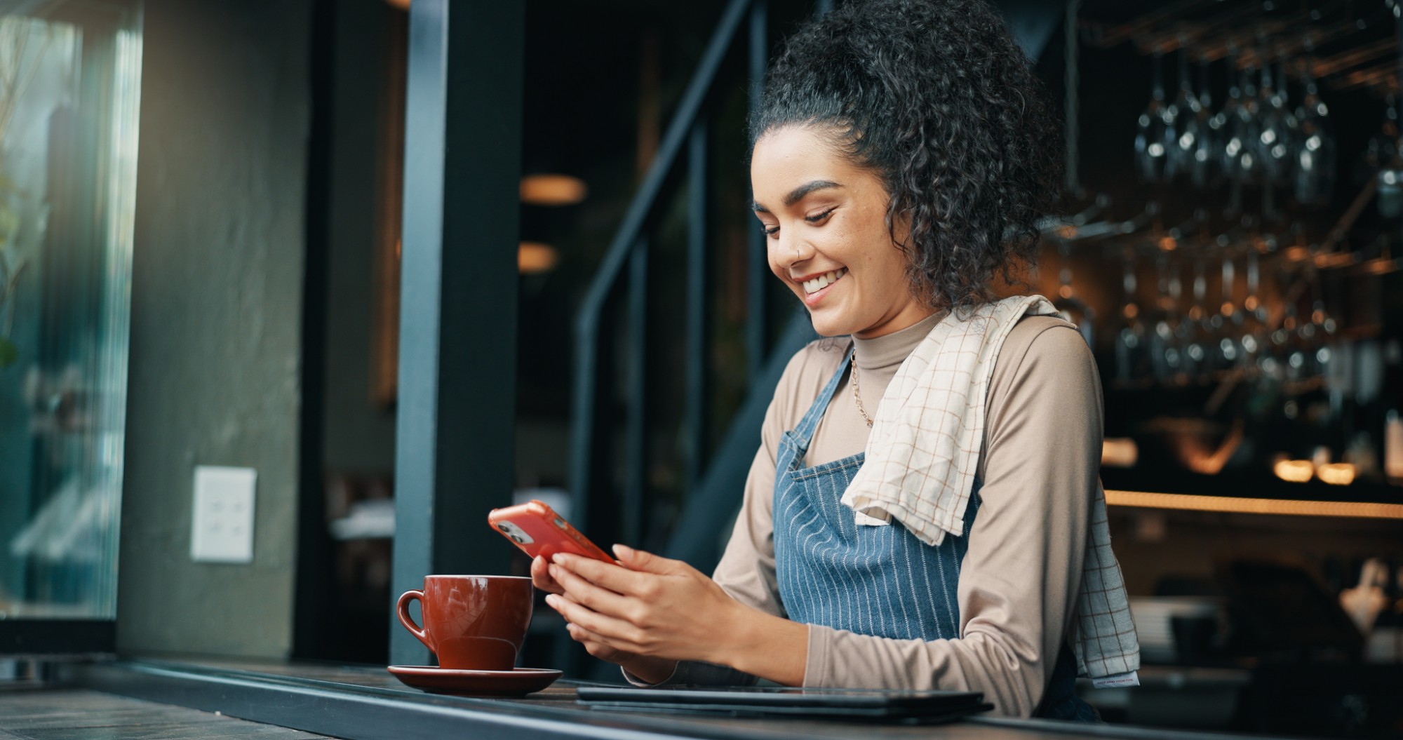 A smiling café worker in a denim apron checking her phone at the counter